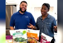 A man and woman stand behind a kitchen counter filled with fruits and vegetables.